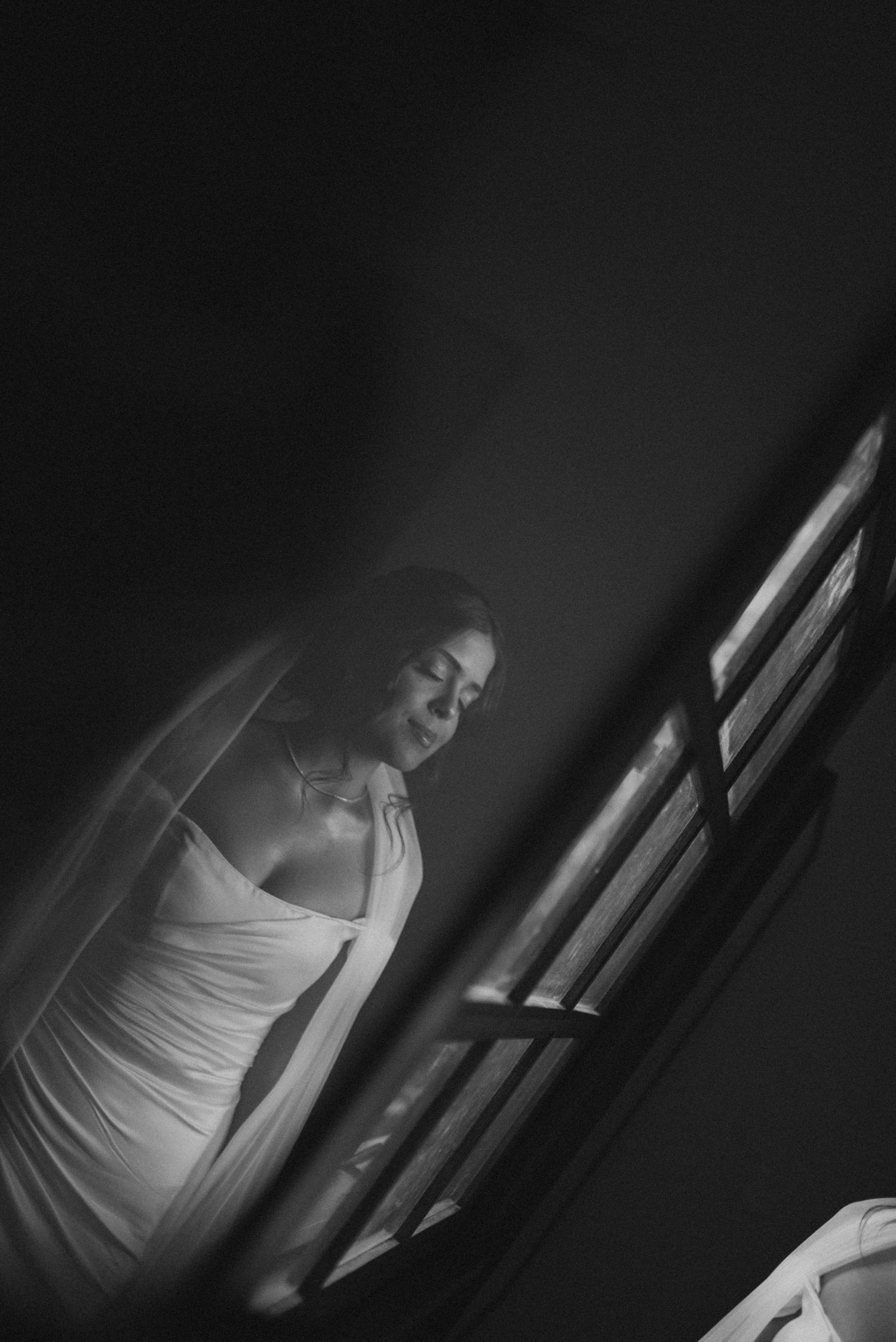 Dramatic black and white photo of person in wedding dress by window with veil flowing.