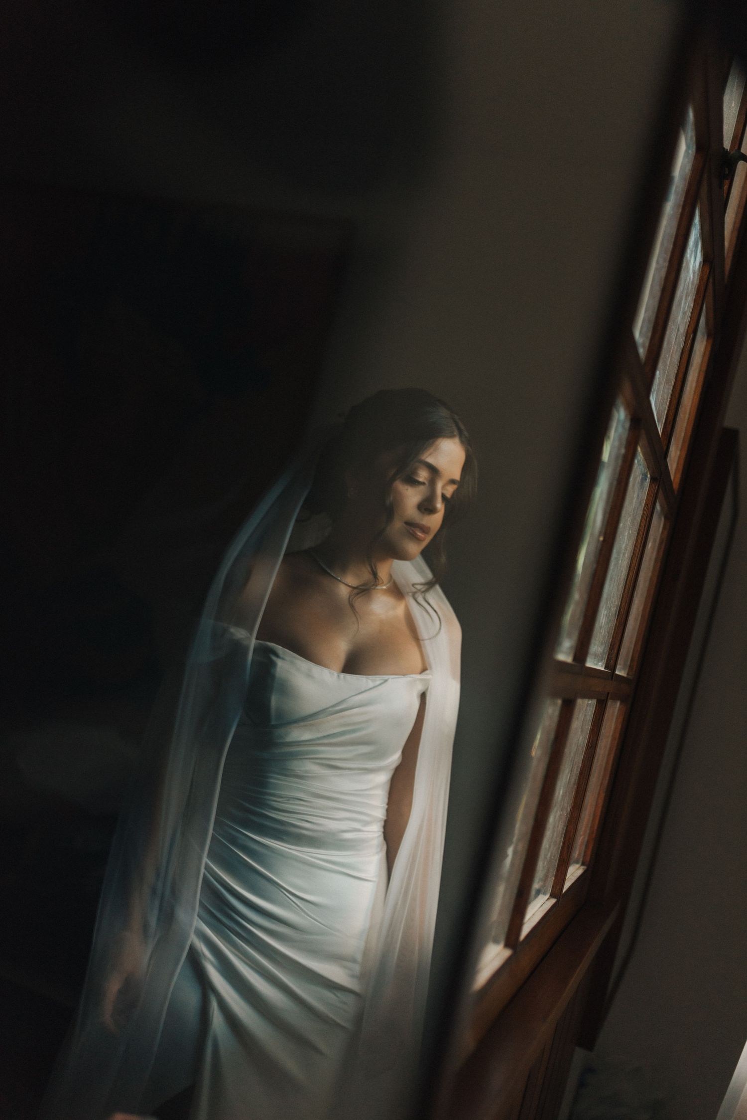 Elegant bridal portrait of a figure in a fitted white gown standing by a window in soft natural light.