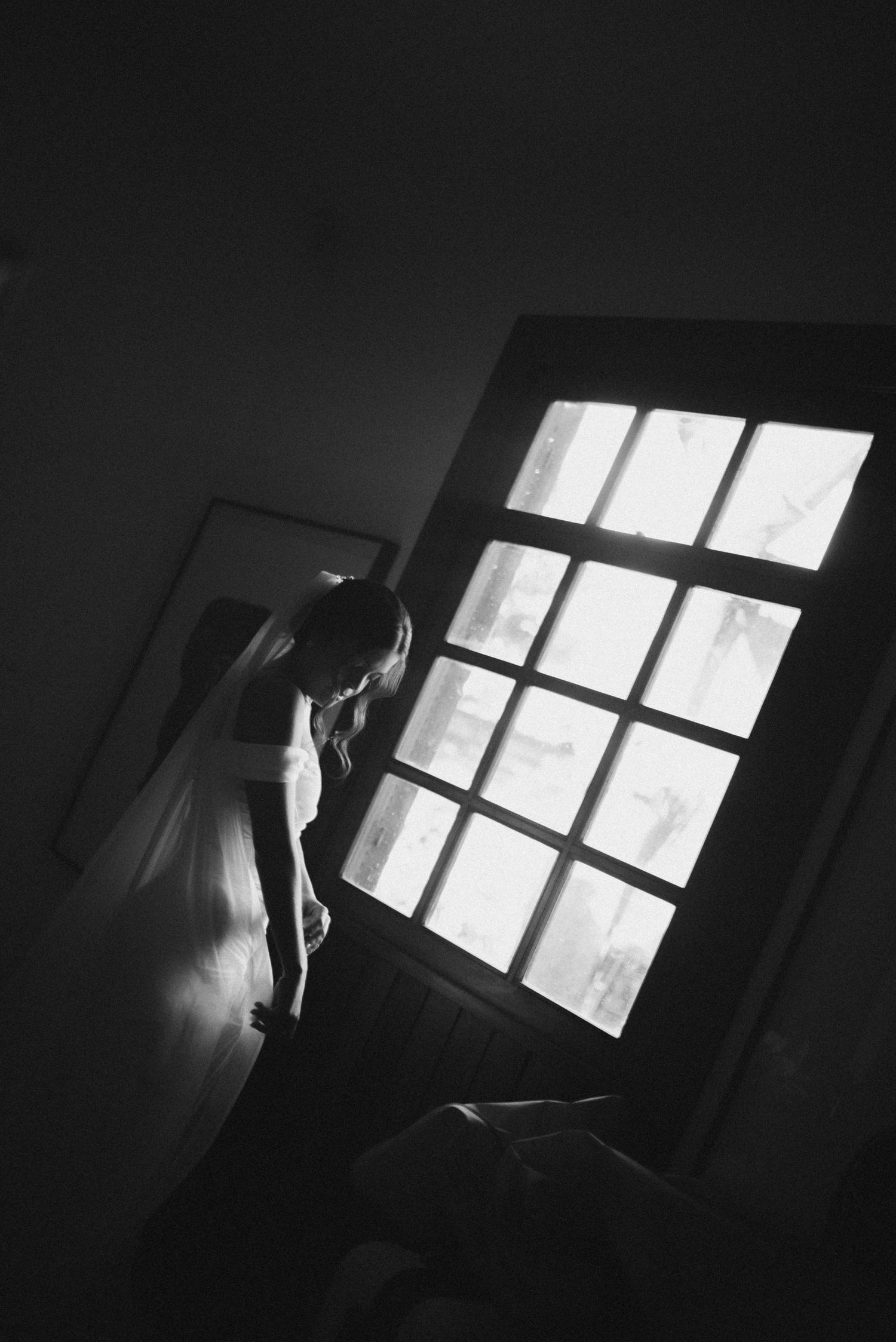 Artistic black and white photo of a bride silhouetted against a bright window with geometric panes.