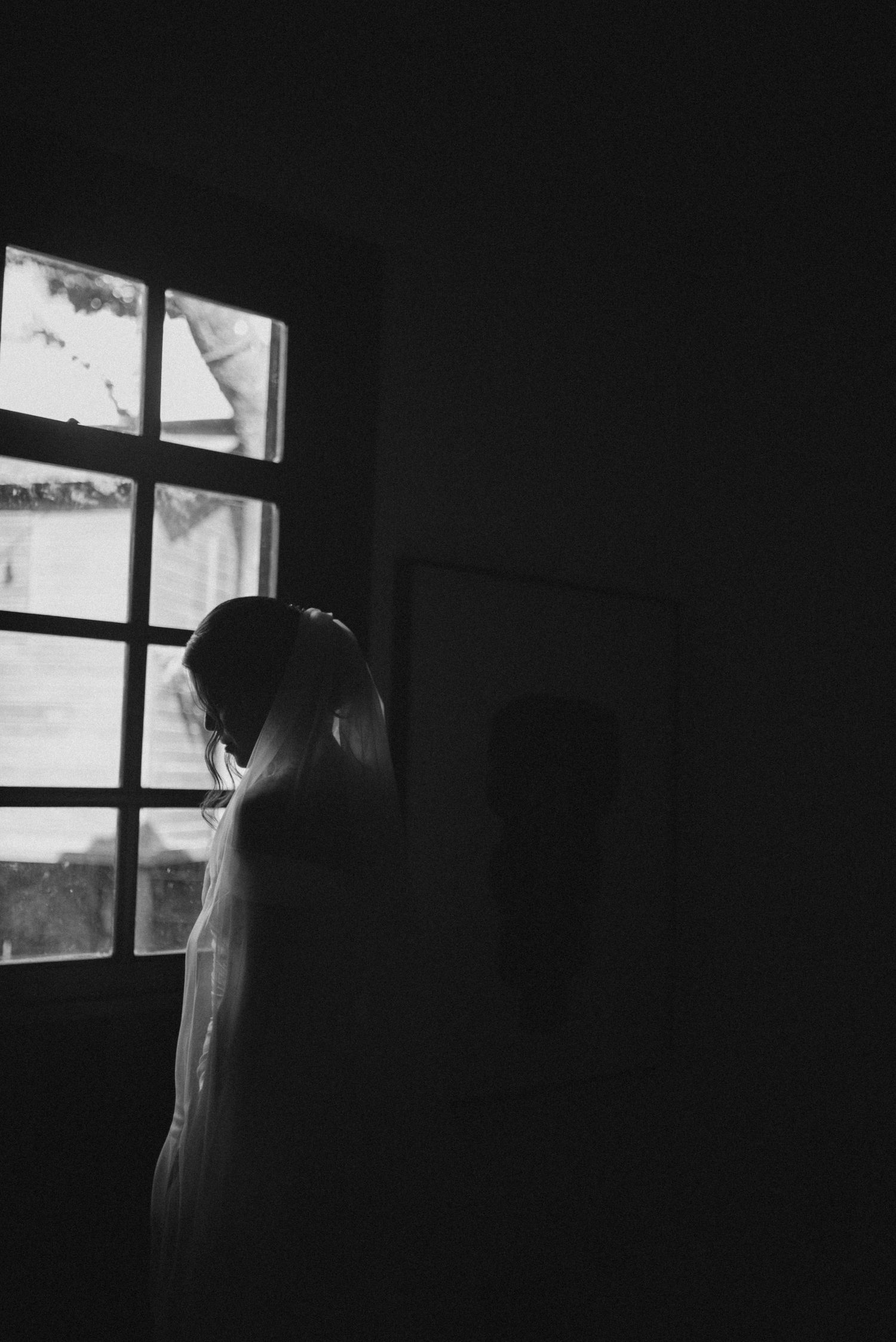 Moody black and white photograph capturing a bridal silhouette against a window.