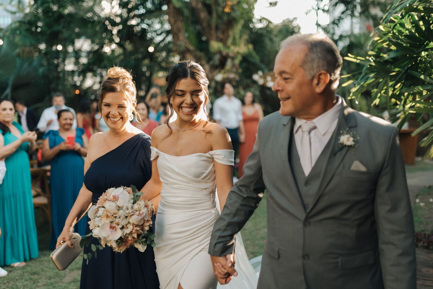 Wedding party walks together through garden path surrounded by guests and tropical foliage.