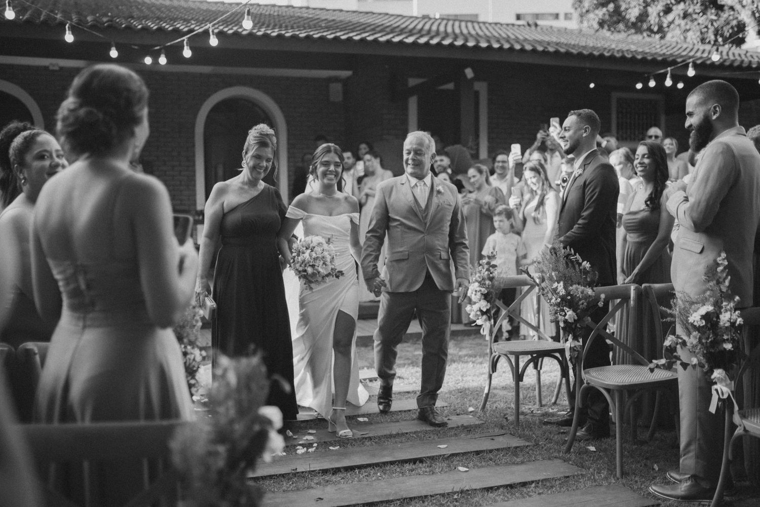 Black and white photo of wedding guests dancing and celebrating at an outdoor reception with string lights.