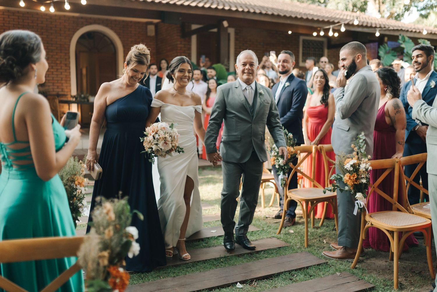 Bride in white dress walks down aisle with attendants in teal and navy dresses at outdoor garden ceremony.