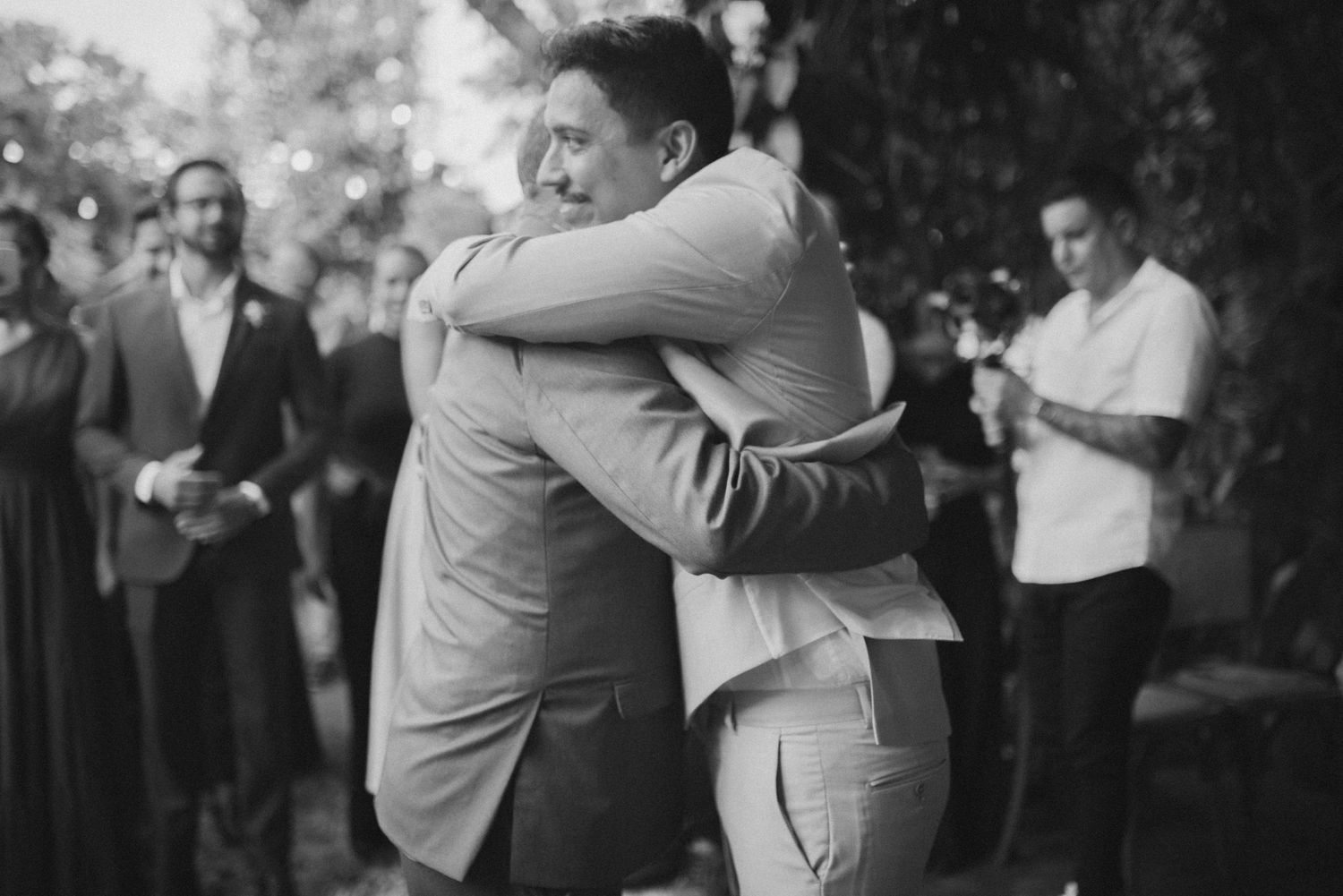 Two people in suits sharing emotional embrace during wedding celebration in black and white.