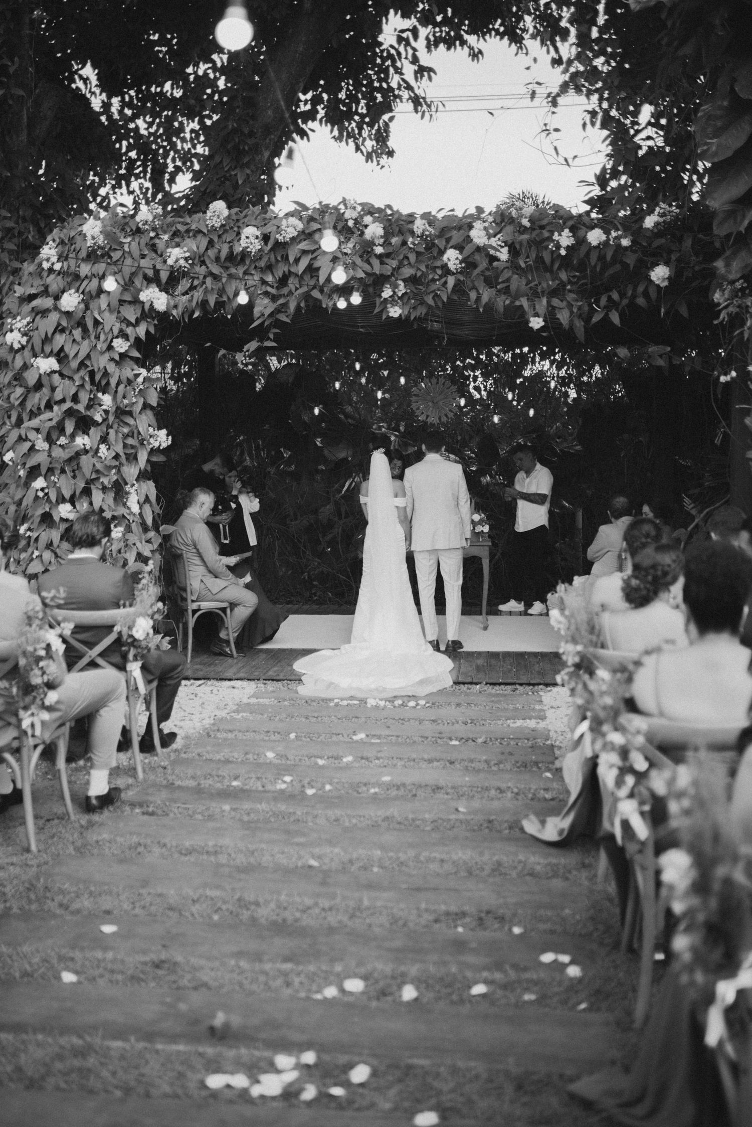 Black and white shot of wedding ceremony under floral arch with guests seated on both sides.