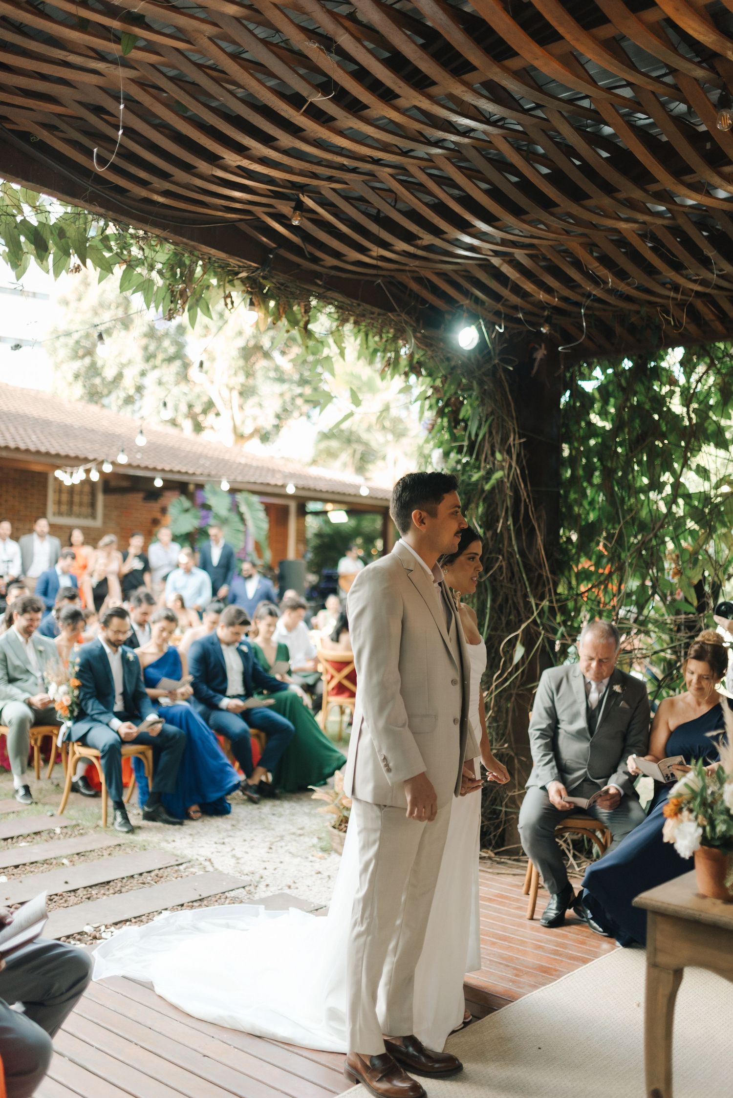 Groom in light suit stands under wooden pergola during outdoor wedding ceremony.