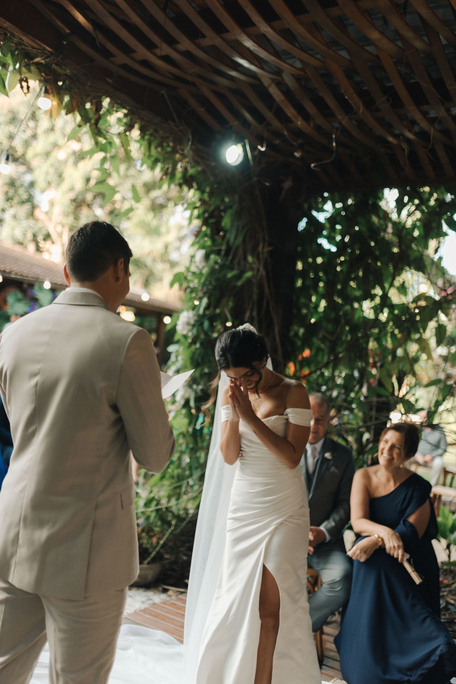 Emotional moment during outdoor wedding ceremony under wooden pergola with guests watching.