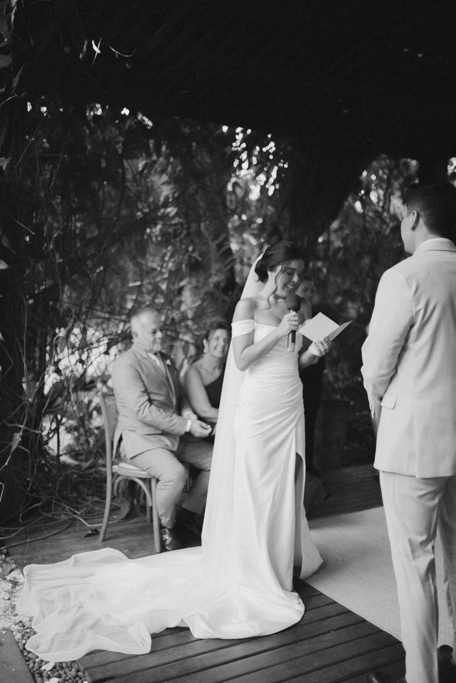 Black and white image of wedding ceremony with bride in flowing dress under natural archway.