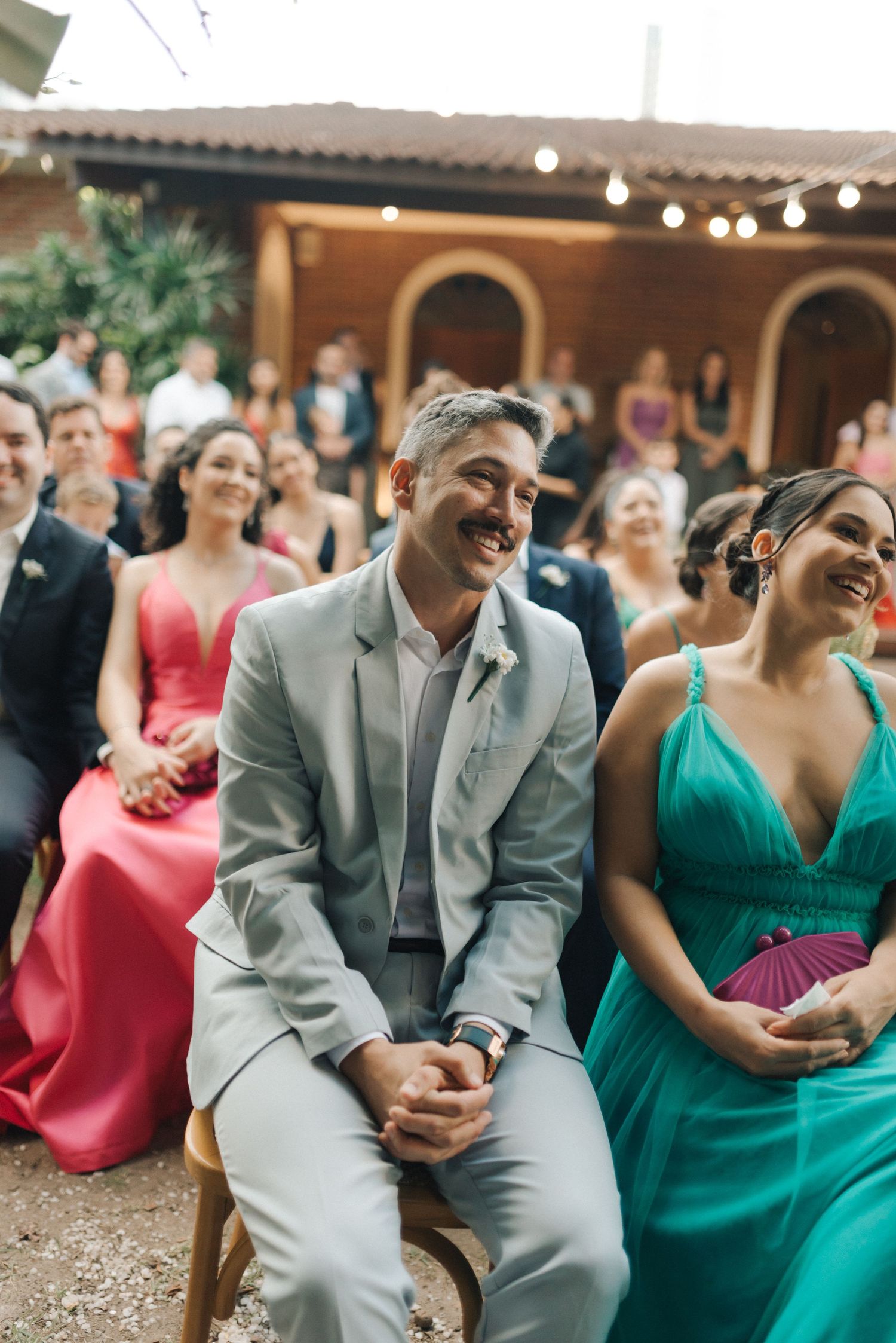 Wedding guests in formal attire smile and enjoy outdoor ceremony with string lights and arches.