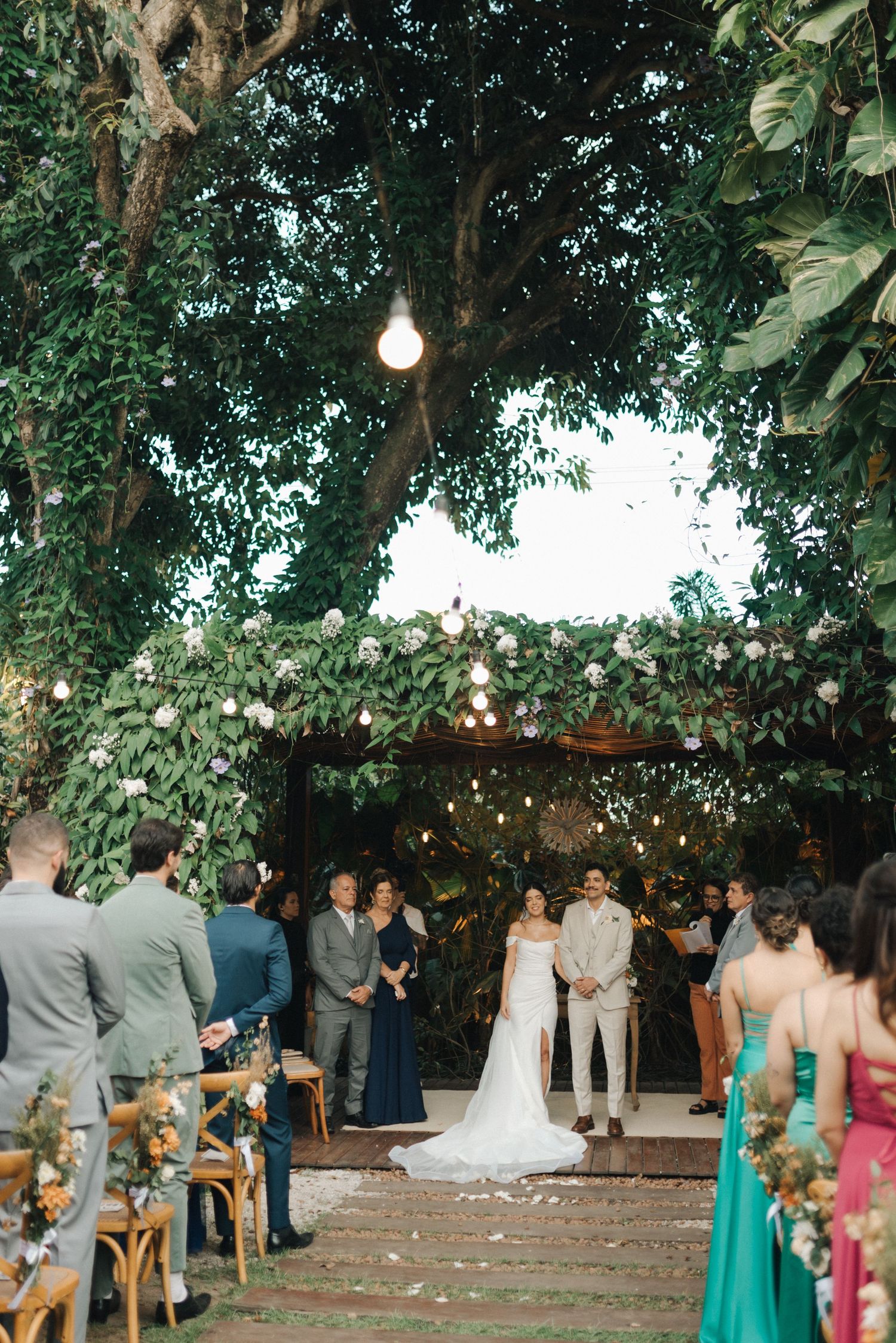 Outdoor wedding ceremony under trees with string lights and floral decorations.