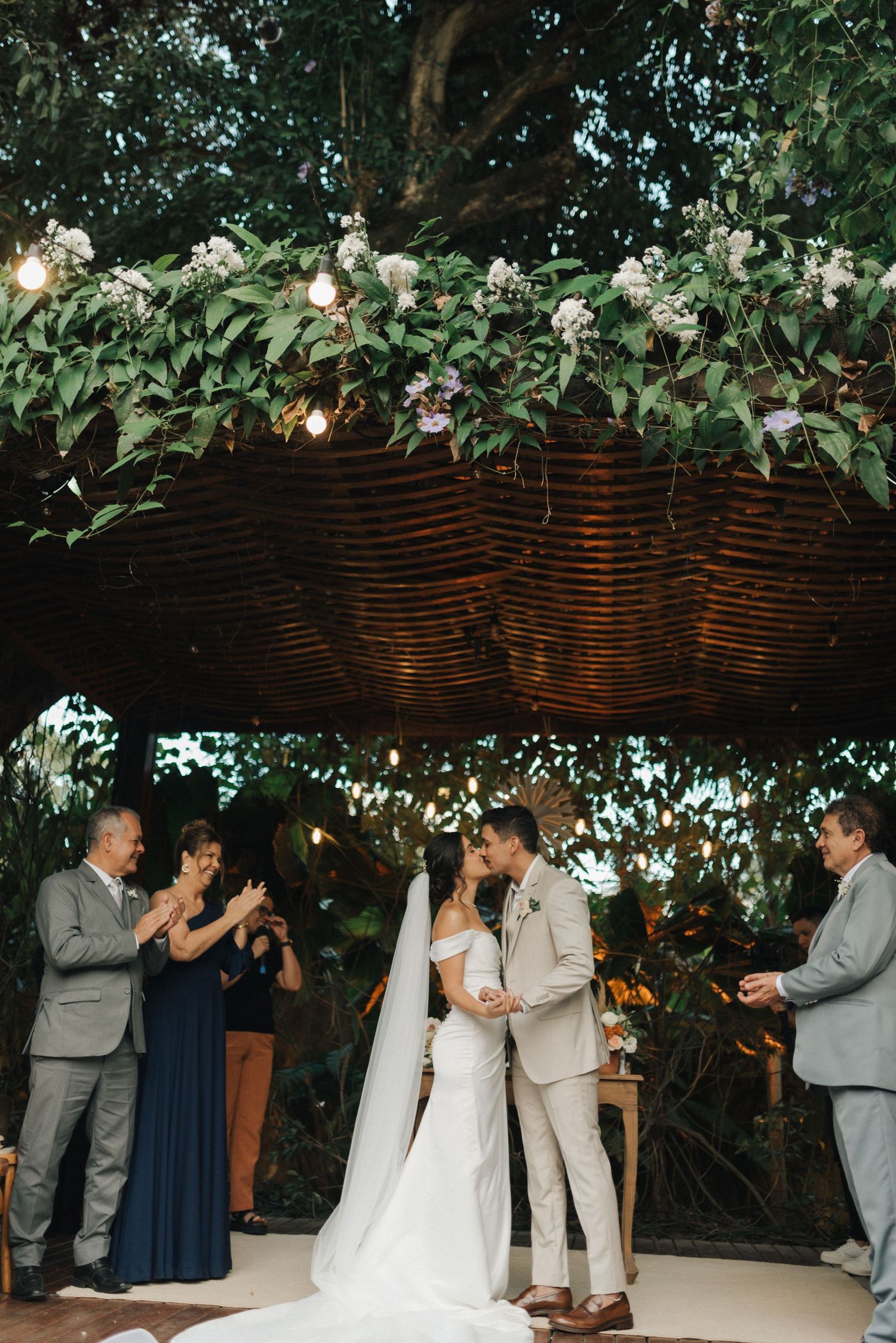 Wedding ceremony taking place under wooden pergola decorated with white flowers and greenery.