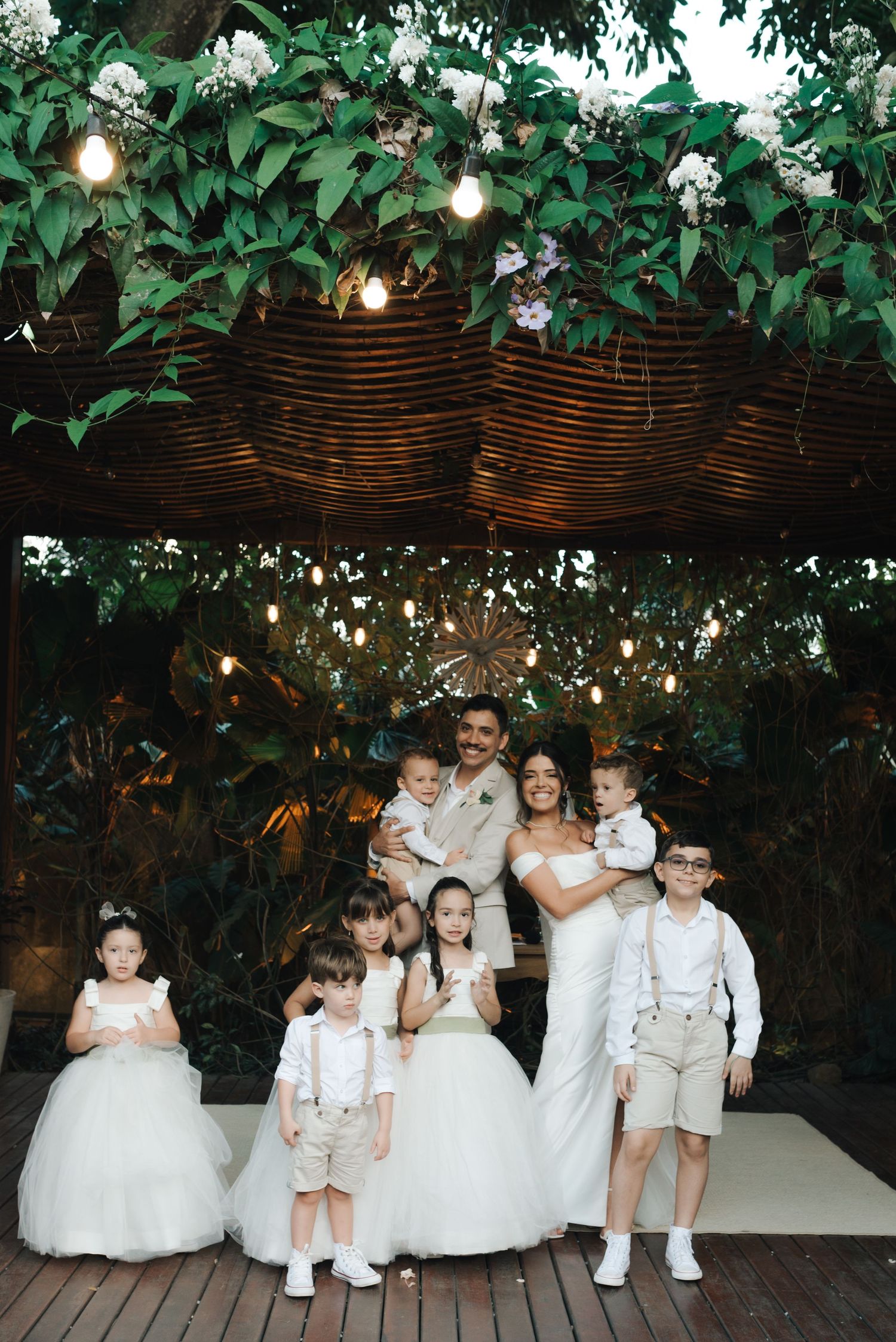 Wedding party photo with flower girls and ring bearers under floral-decorated pergola.