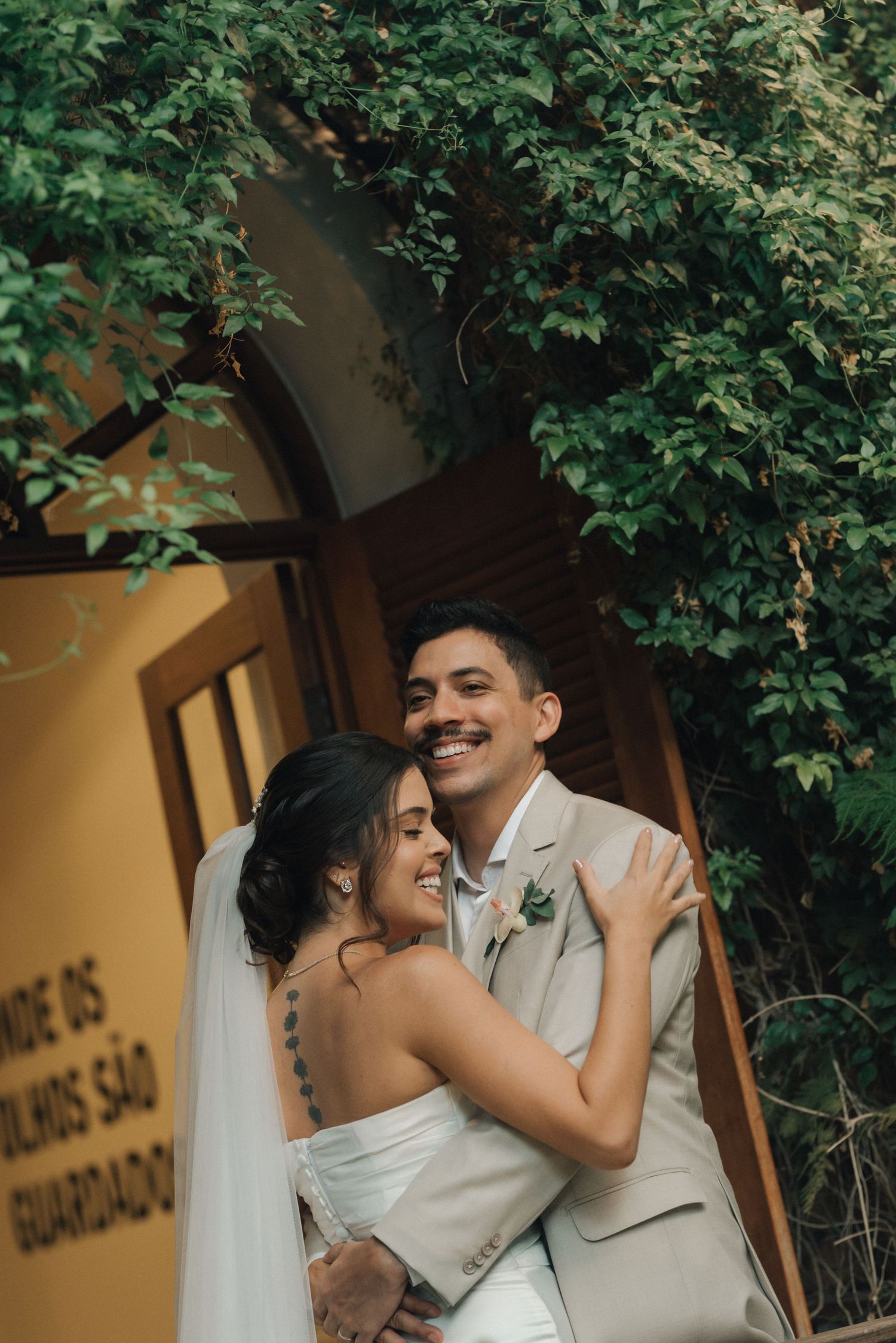 Happy wedding couple embracing under leafy greenery and arched doorway.