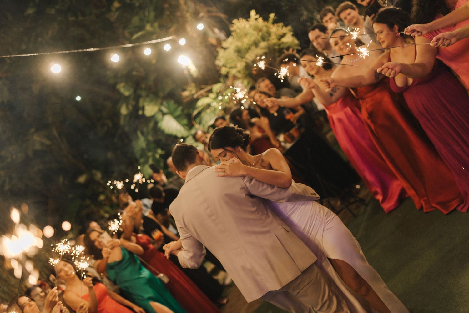 Wedding party and guests celebrate under twinkling lights in an outdoor evening reception.