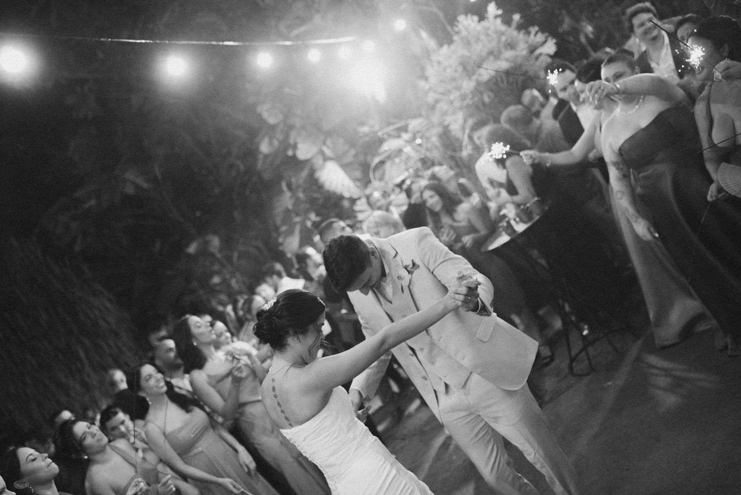 Black and white photo of newlyweds dancing surrounded by celebration guests with sparklers.