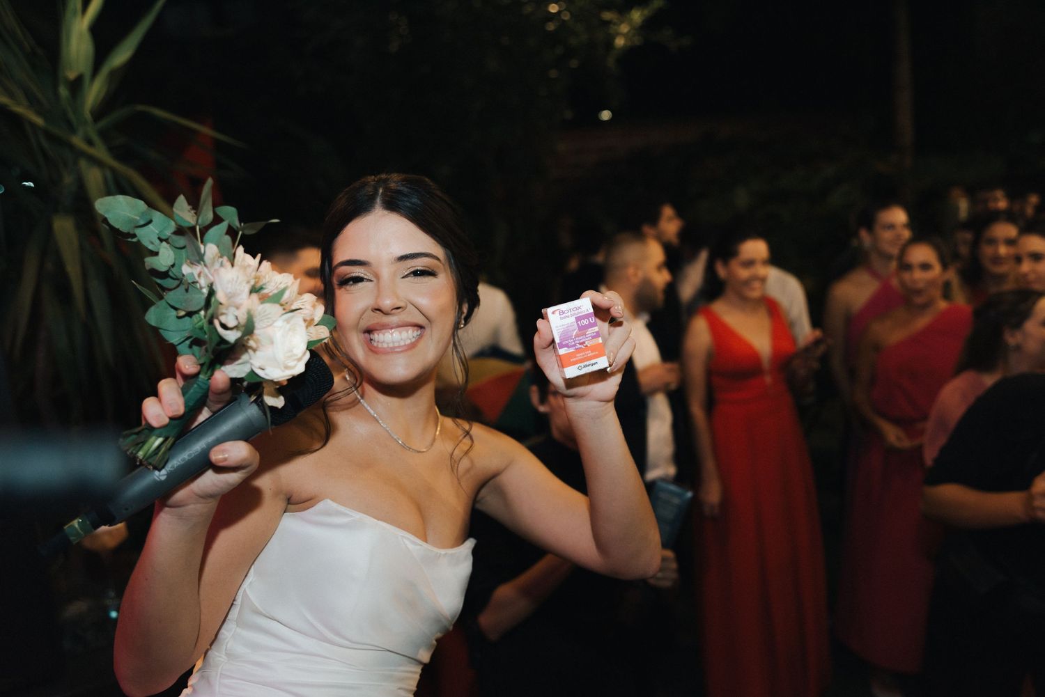 A bride celebrating with flowers and guests at an outdoor evening reception.