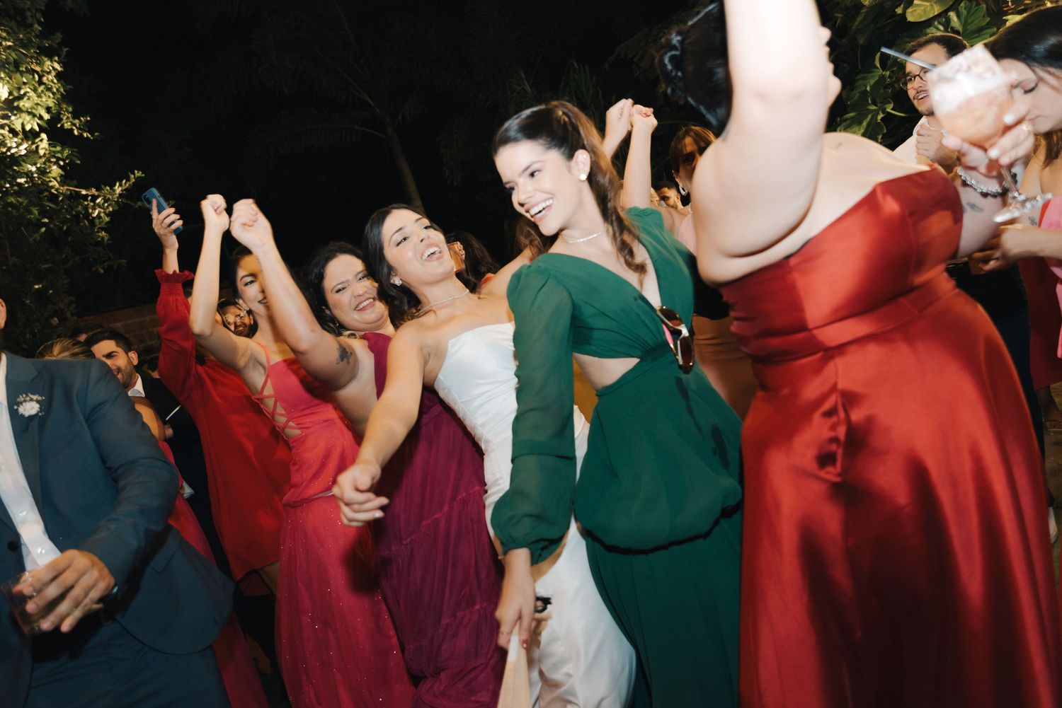 Celebratory group dancing in red and green formal dresses at a nighttime event.
