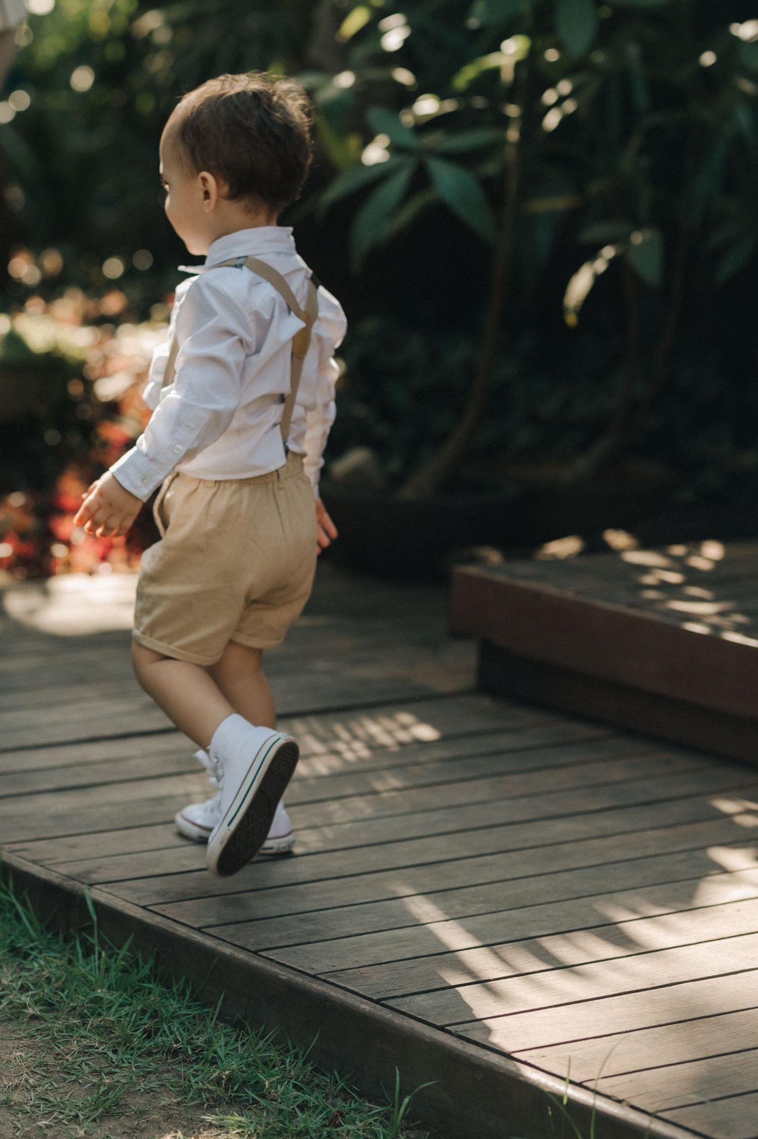 Young boy in formal attire with suspenders walking on wooden deck in garden setting.