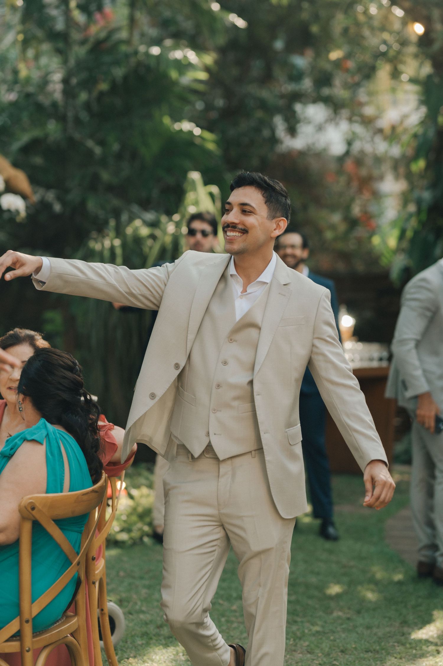 Groom in beige suit walking through garden reception area with joyful expression.