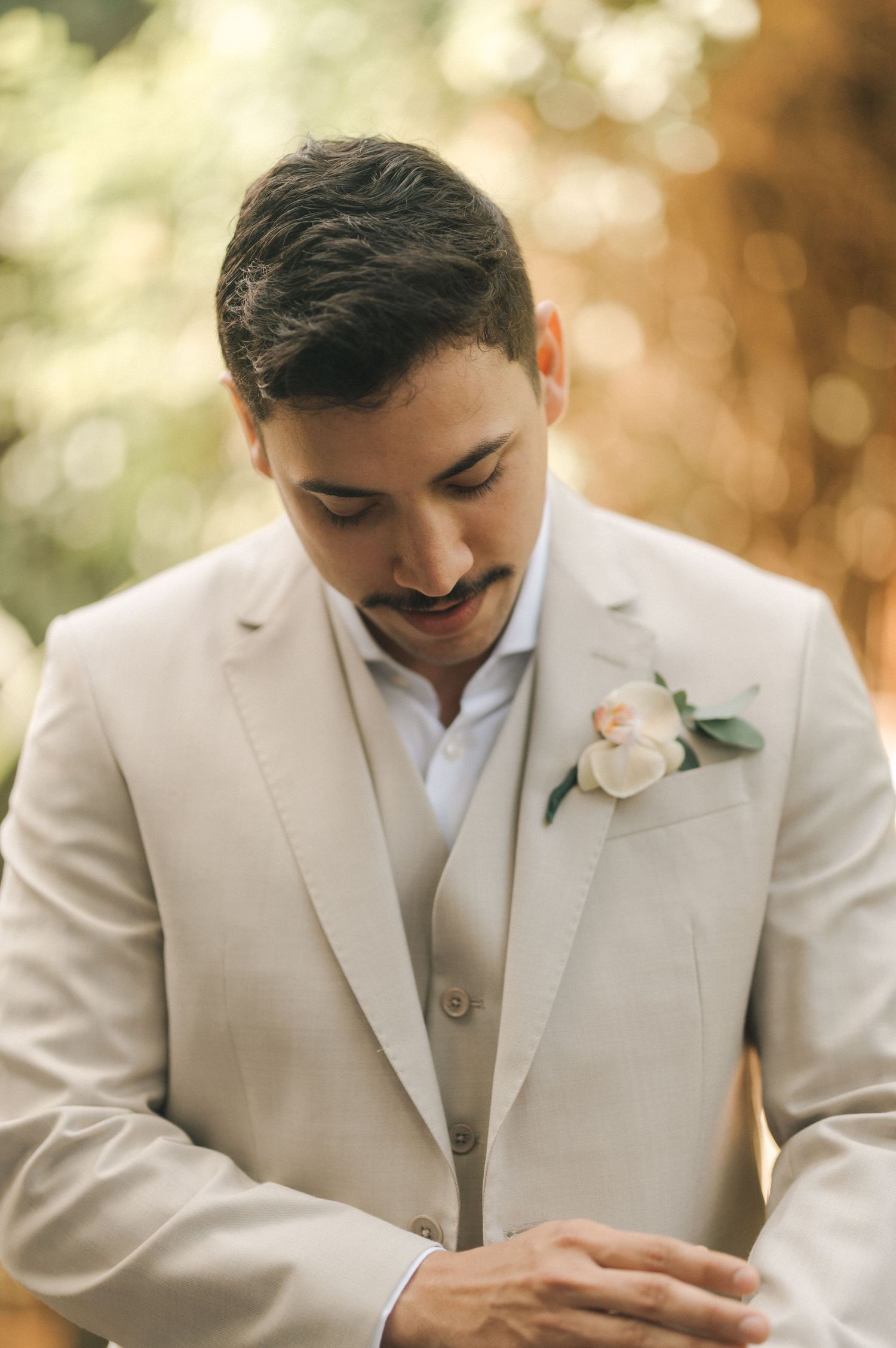 Close-up of groom in light beige suit with white boutonniere looking down.