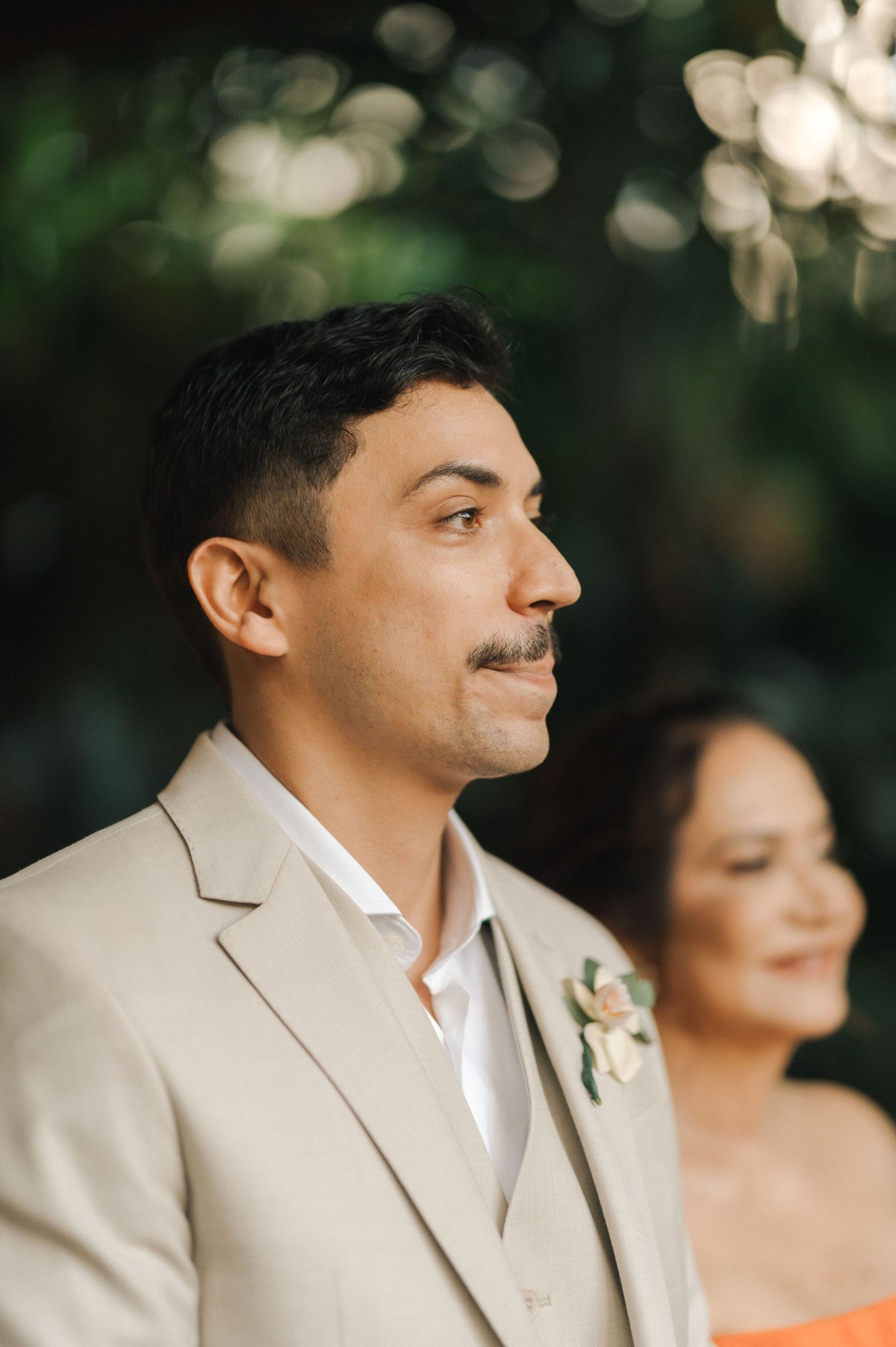 A groom in a beige suit smiling during an outdoor wedding ceremony.