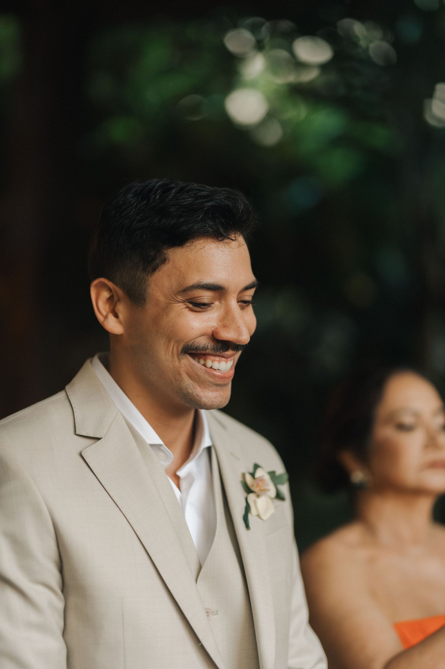 Groom in beige suit smiling during outdoor wedding ceremony.
