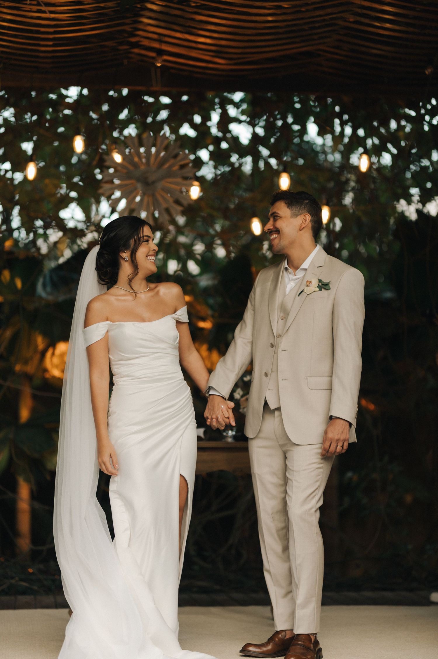 Bride in elegant white gown and groom in beige suit hold hands during their outdoor ceremony.