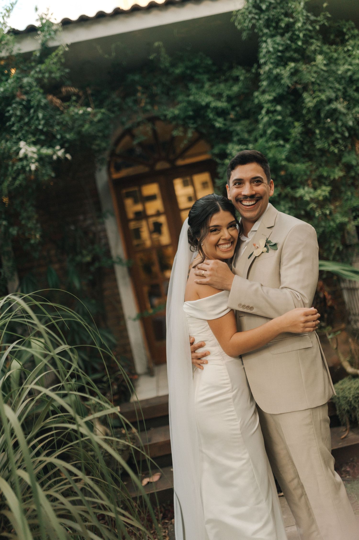 A newlywed couple embraces and smiles in front of an archway surrounded by greenery.