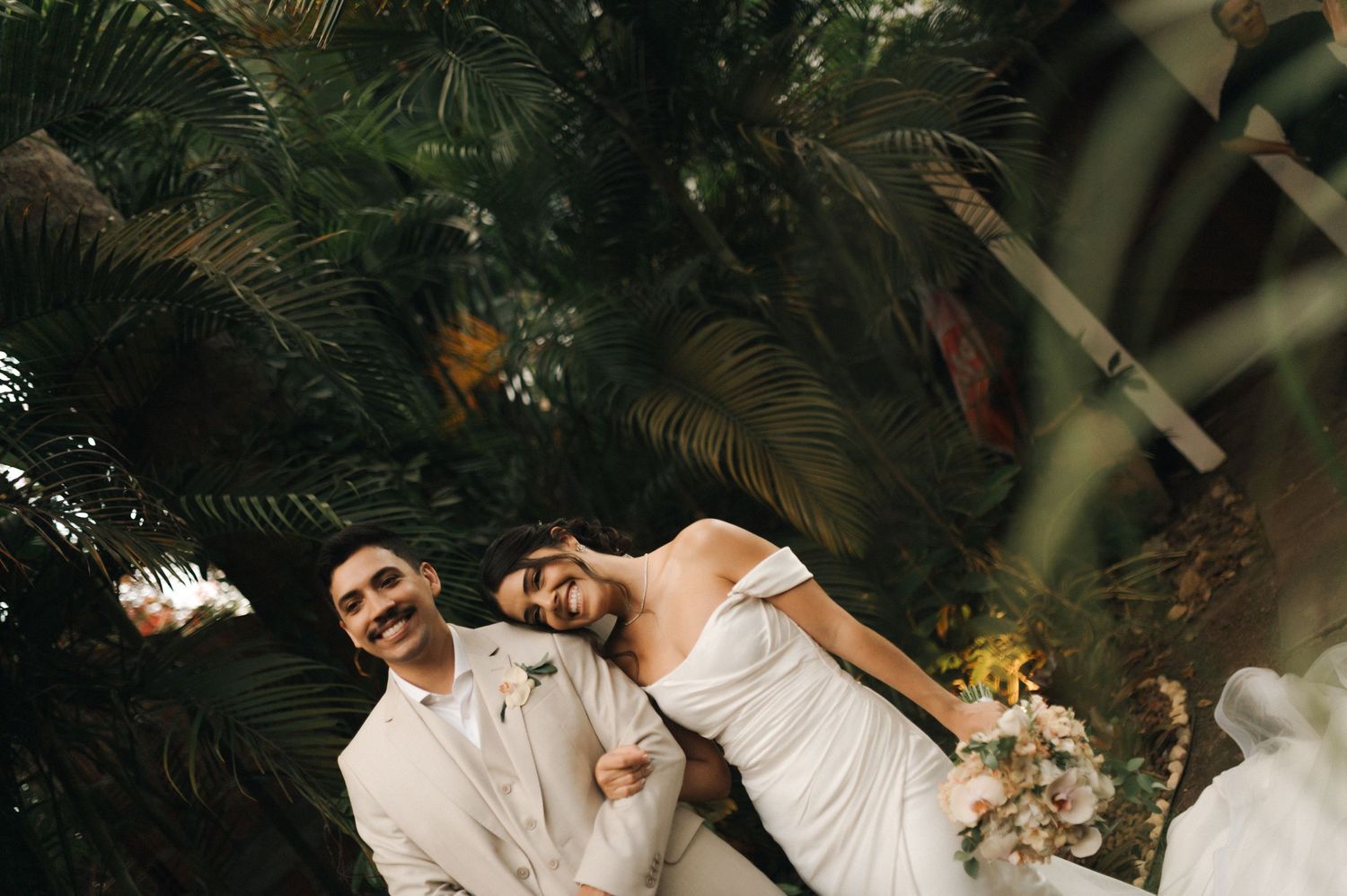 A joyful wedding couple laughing together against a backdrop of lush palm trees in a tropical setting.