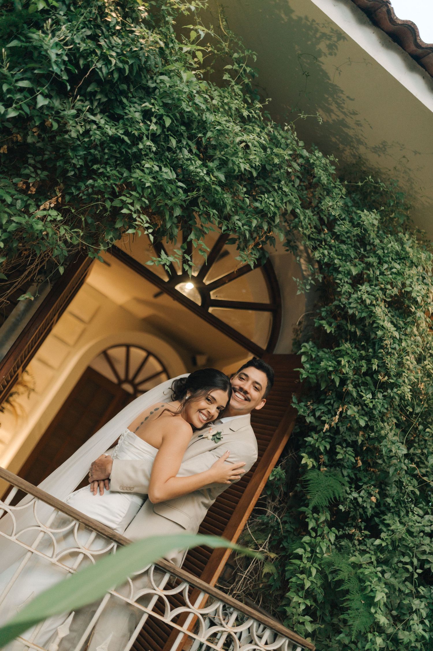A romantic embrace captured on a balcony with lush greenery and an arched doorway.
