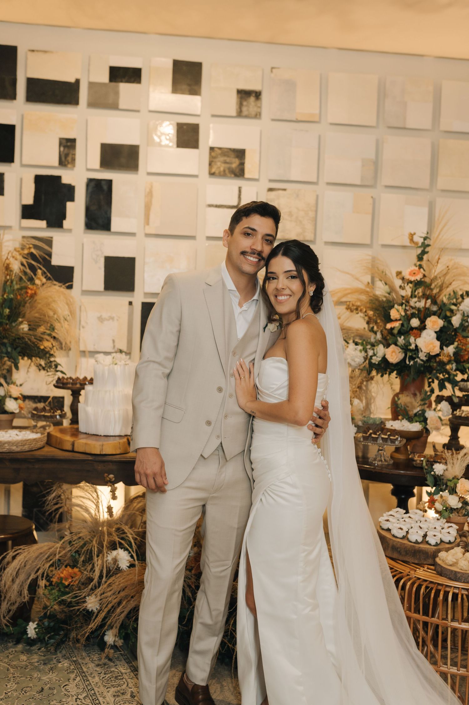 A wedding couple poses in front of a decorative wall display with floral arrangements.
