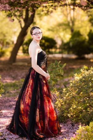 Person in formal gown poses in garden setting with autumn colors in background.