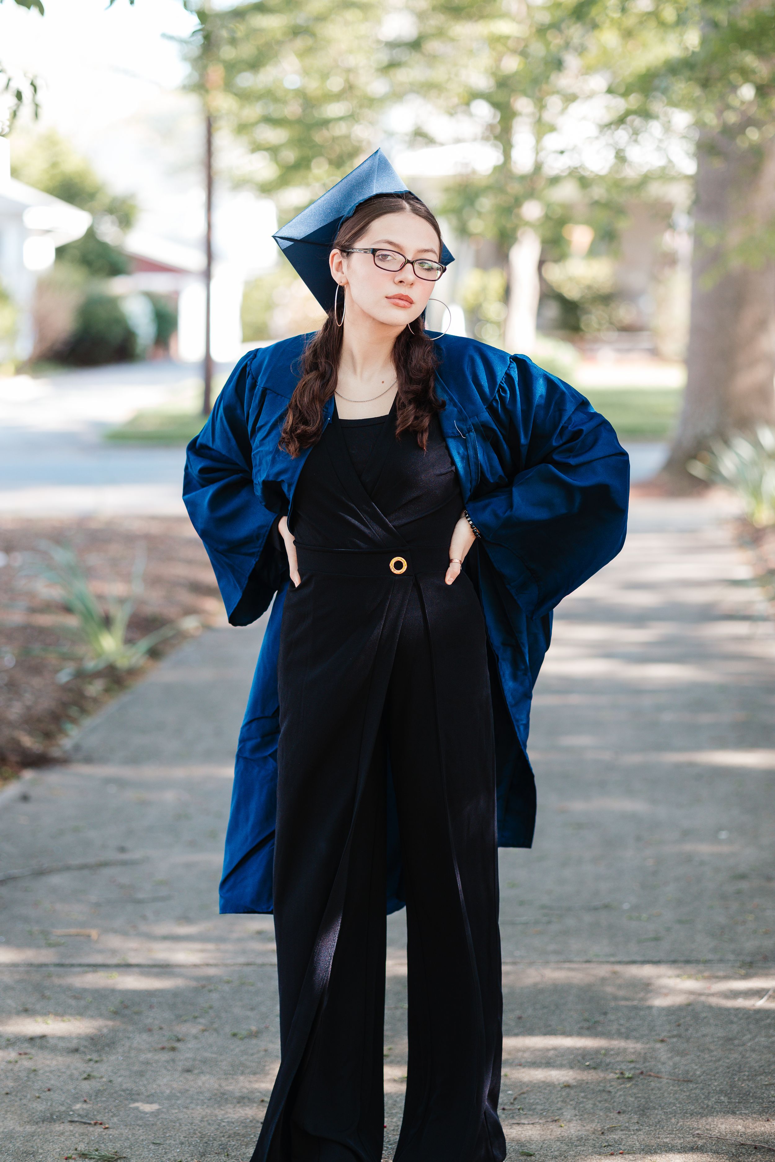 Graduate in blue academic regalia and black outfit poses on campus walkway.