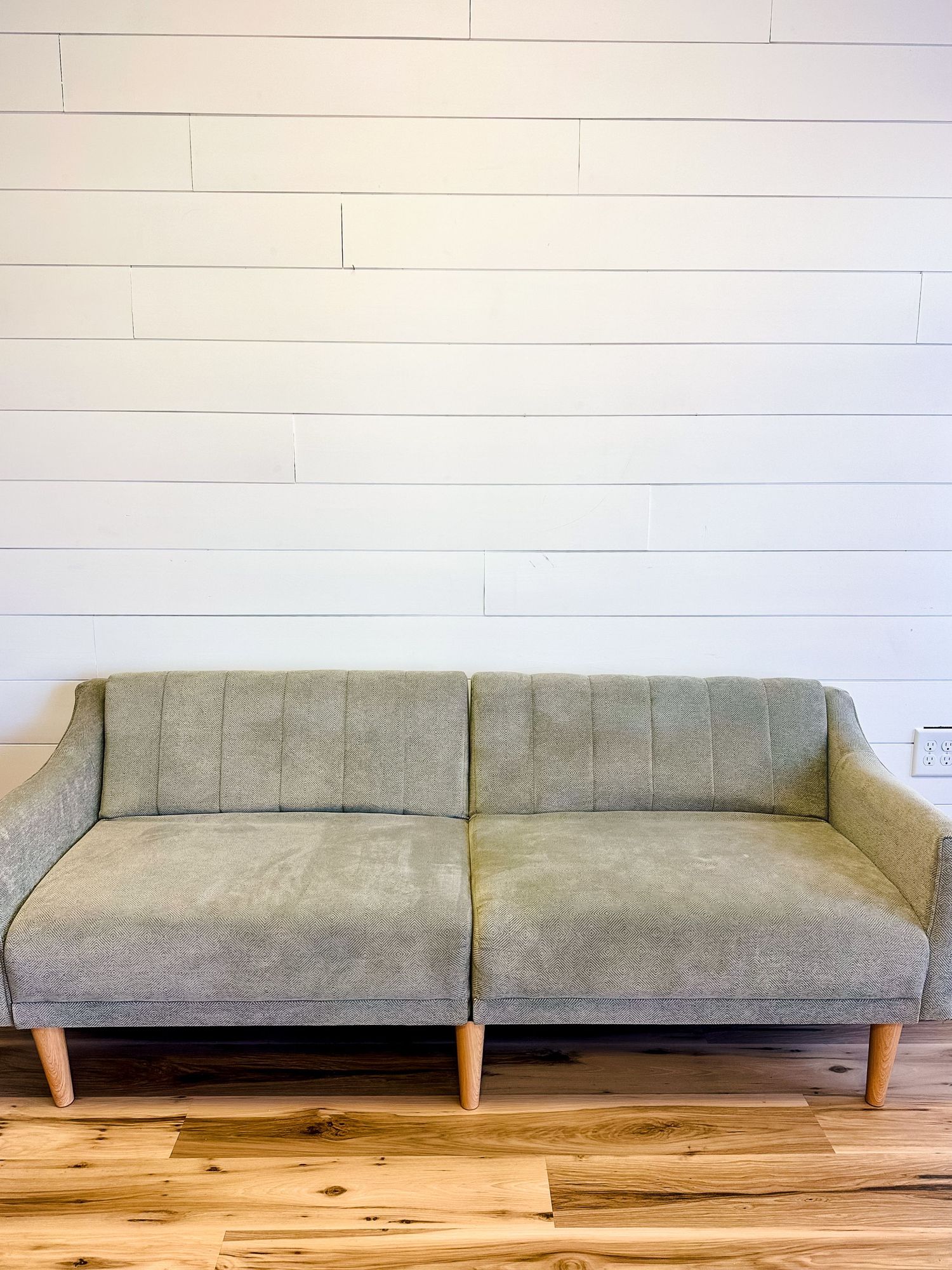Modern grey upholstered sofa with wooden legs against white shiplap wall on hardwood flooring.