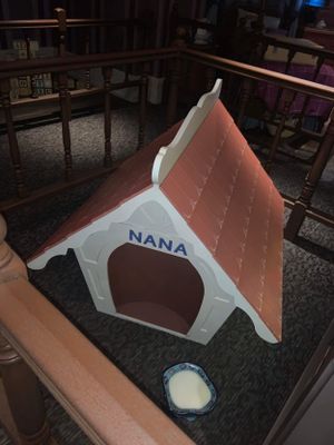 A pink and white dog house labeled 'NANA' with a peaked roof and rounded entrance, sitting indoors with a water bowl nearby.