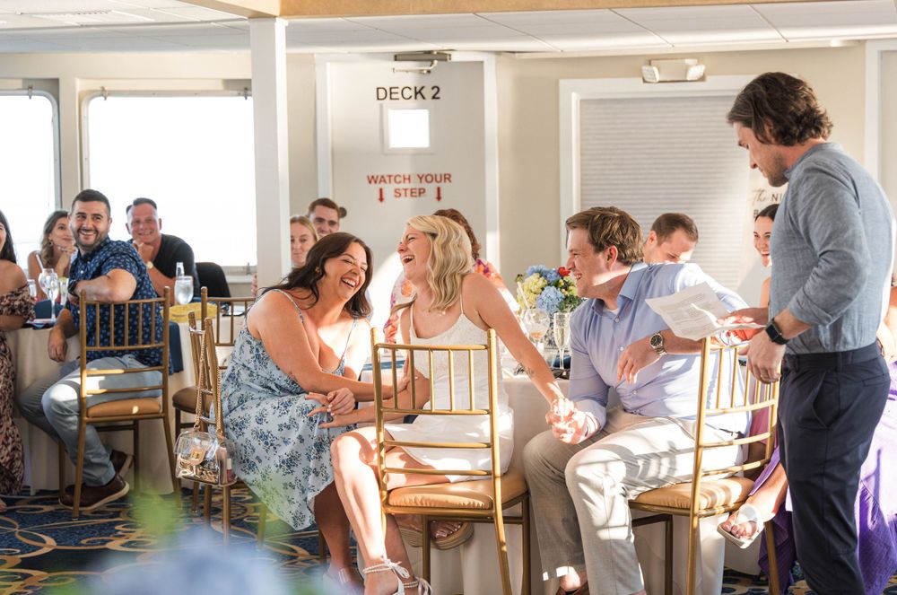 Guests celebrating and laughing during wedding rehearsal dinner on a boat in Gloucester, MA, with sunset in the background.