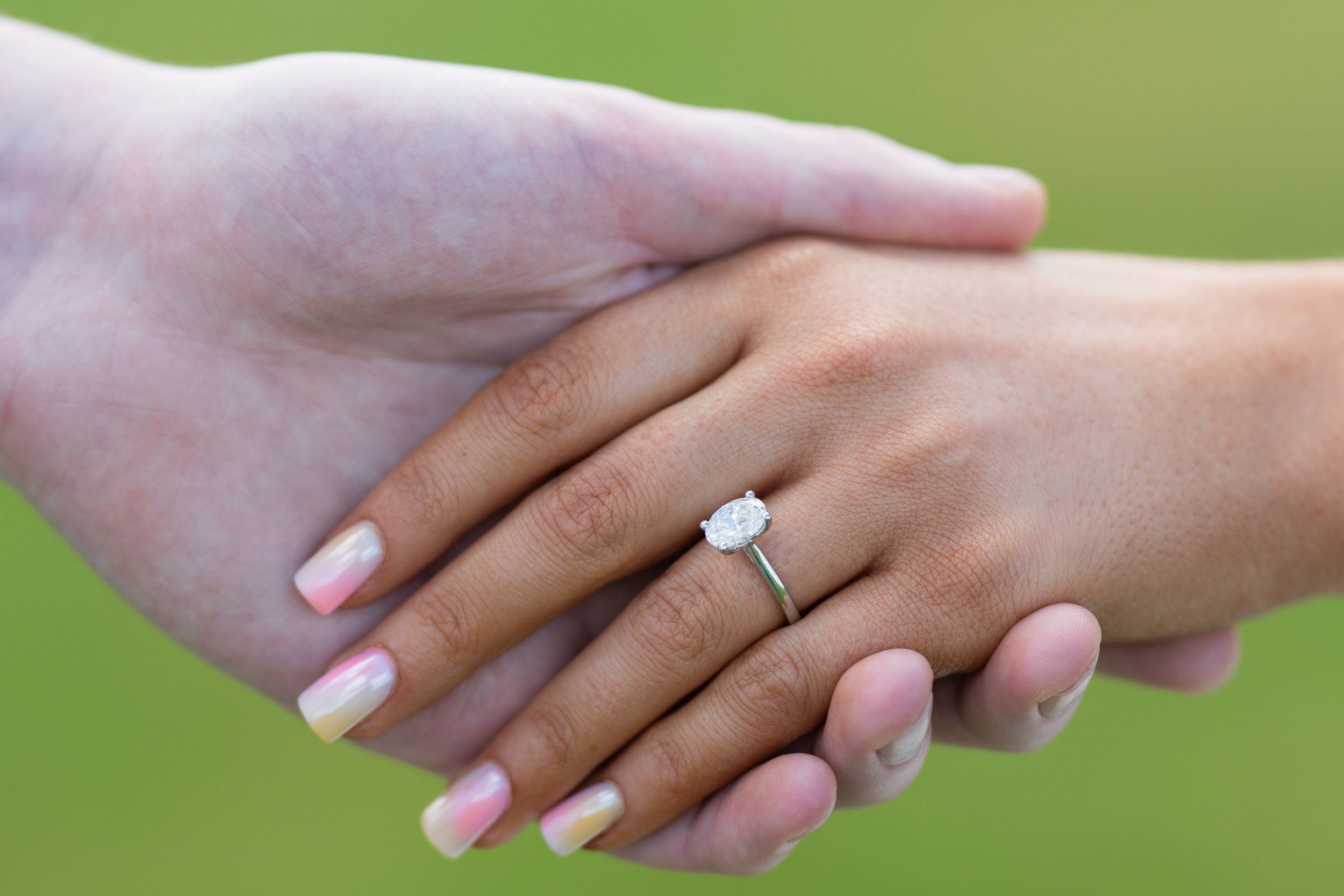 Close-up of hands displaying an elegant engagement ring against a green background.