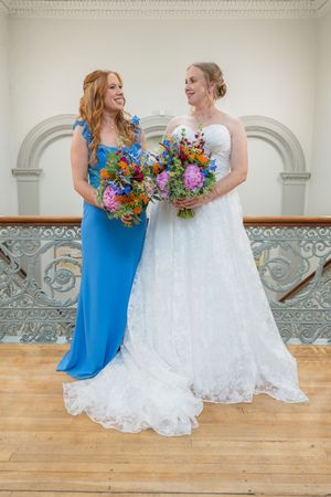 Two people in blue and white dresses share a joyful moment with colorful bouquets in an elegant indoor setting.