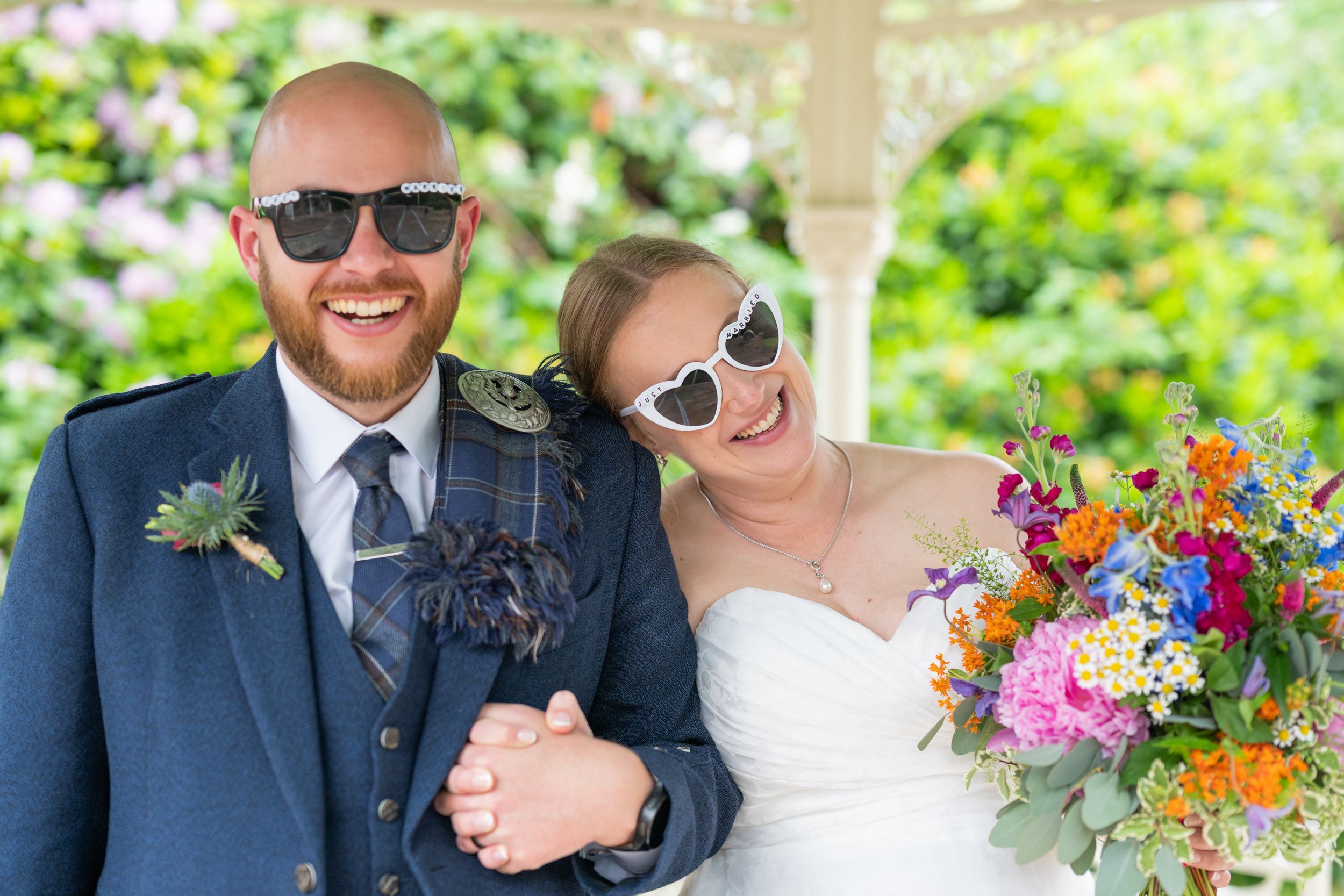A couple wearing sunglasses shares a happy moment outdoors with vibrant bouquets.