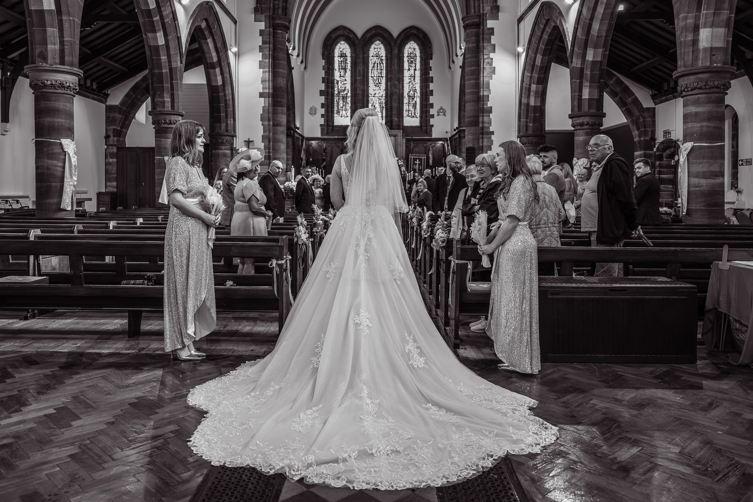 Black and white photo of a bride in a long lace gown and veil standing at the church altar with bridesmaids.