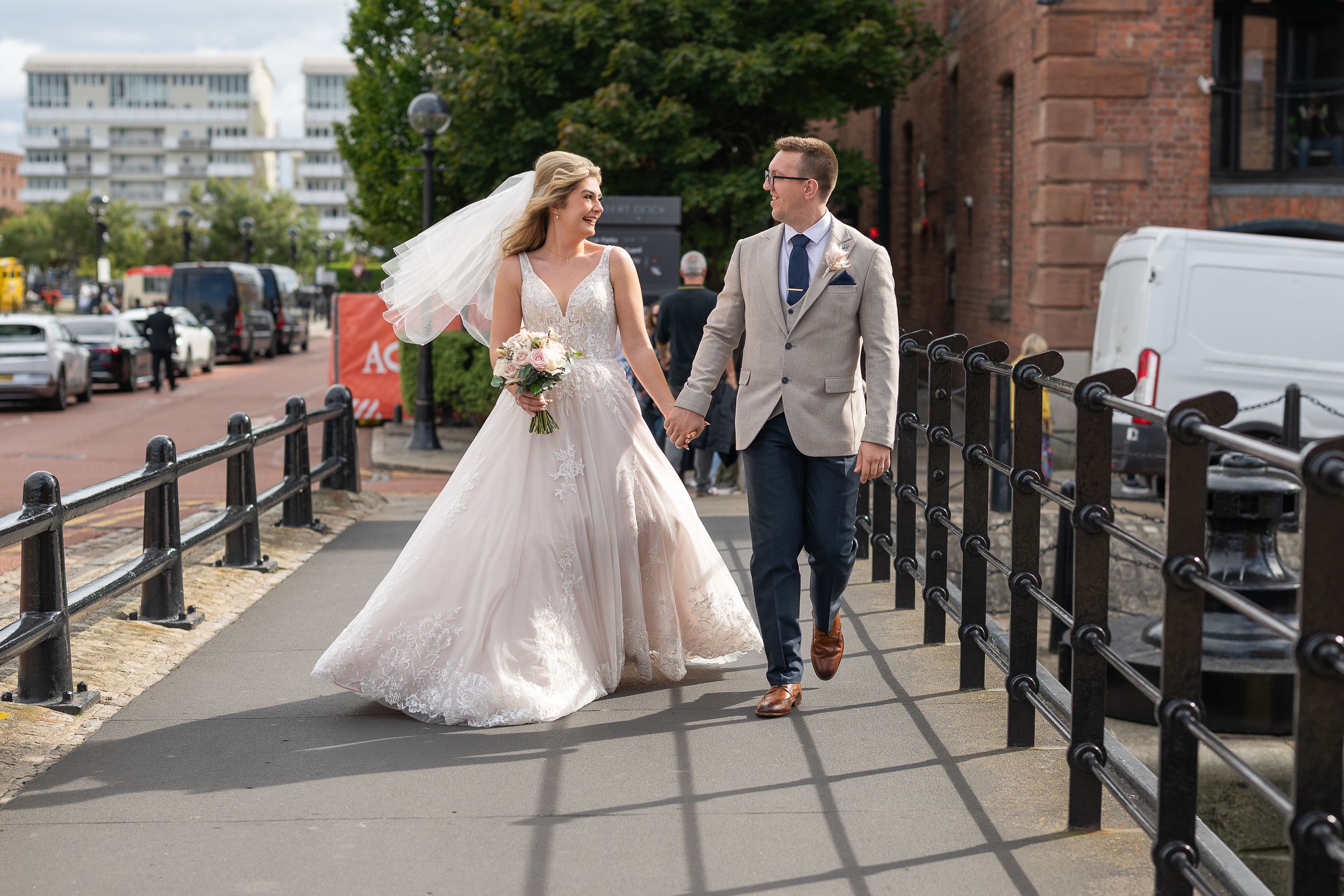 Bride and groom walk hand in hand on a sunny day, bride in white gown with veil, groom in grey suit.