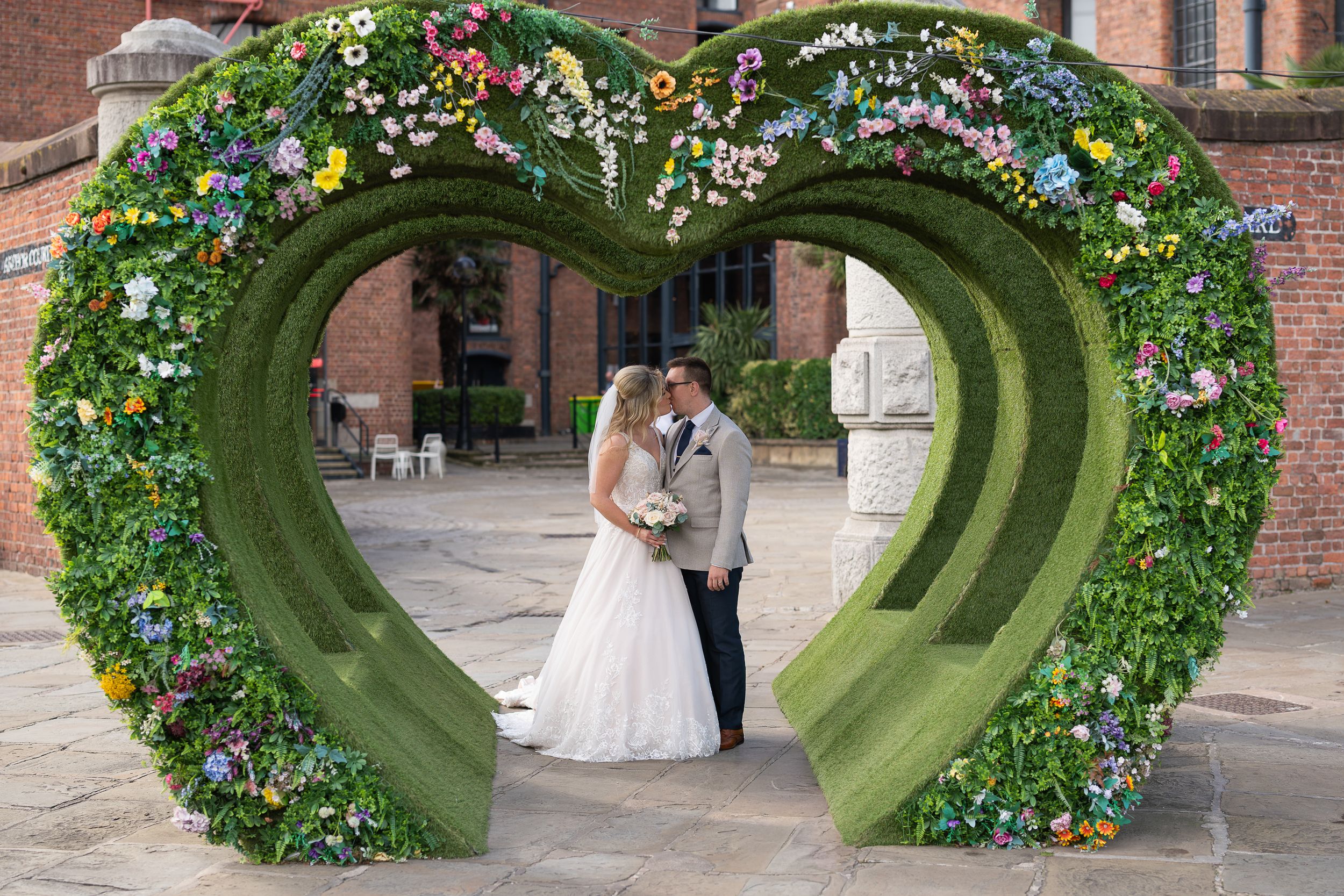 Bride and groom kissing under a heart-shaped floral arch with colorful blooms at an elegant wedding venue.