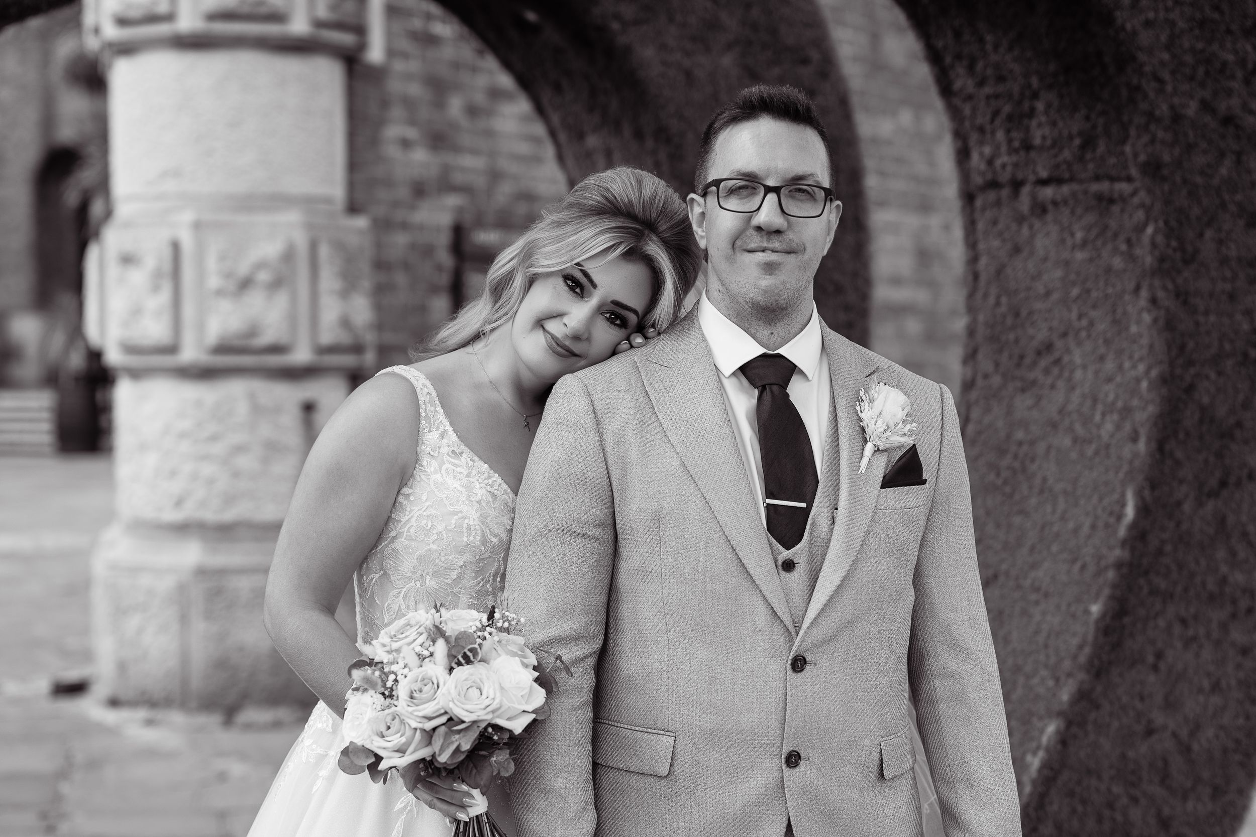 Black and white wedding photo of smiling bride with bouquet leaning on groom in suit near stone arches.