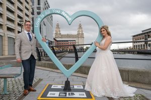 Bride and groom posing by a teal heart sculpture reading Love Is All You Need at Liverpool waterfront.