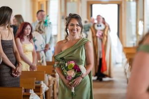 Smiling bridesmaid in green one-shoulder dress walks down church aisle holding pink bouquet during wedding ceremony.