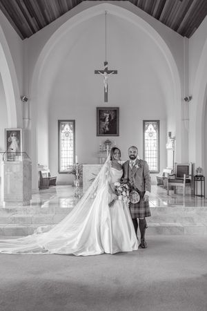 Black and white photo of a bride and groom posing inside a church, with a crucifix and arched ceiling behind them.