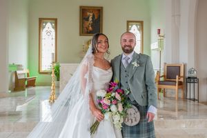 Bride in white gown and groom in gray kilt pose together inside a decorated church after wedding.