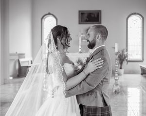 Black and white wedding photo of bride in veil and groom in kilt sharing a joyful moment in a church.