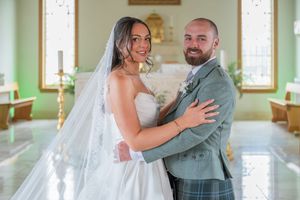 Bride in white gown and veil embraces groom in tartan kilt inside a green-walled chapel on their wedding day.