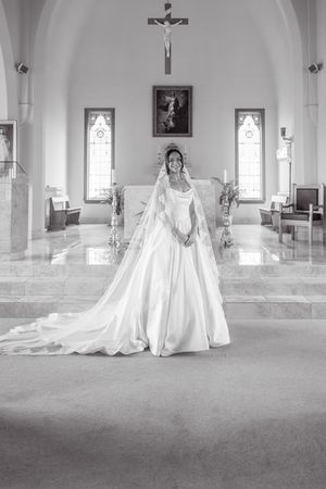 Bride in elegant white ball gown and veil stands at church altar with crucifix, in black and white photo.